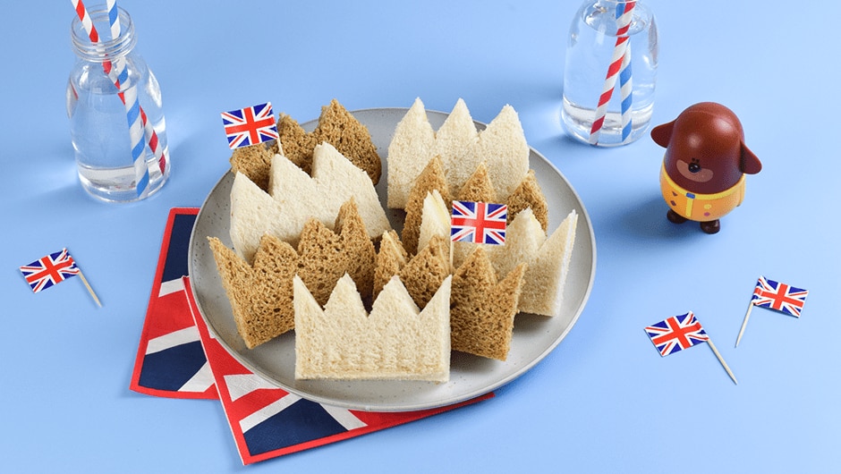 A lifestyle image of a plate of sandwiches shaped like crowns decorated with Union Jack mini flags. In the background are more mini flags, some Union Jack napkins, a Duggee figurine and two glass bottles filled with water and finished with blue and red straws inside.