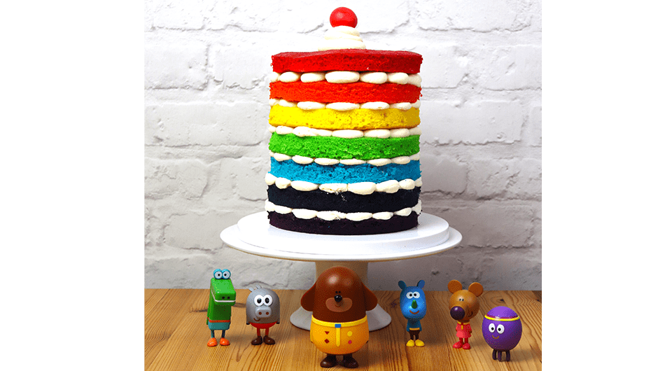 Completed rainbow cake placed on a cake stand with Hey Duggee figurines in front.