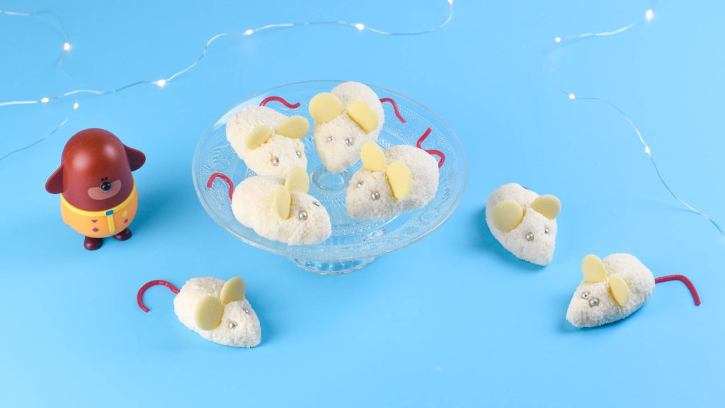 Lifestyle image of several coconut mice, some on a desert stand others on a blue counter top with a Duggee figurine and some fairy lights.